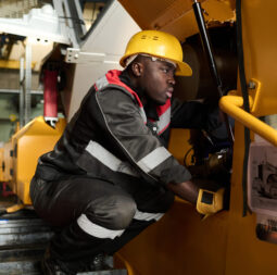 Young professional repairman or mechanic checking mechanism of industrial machine while sitting on squats in factory workshop